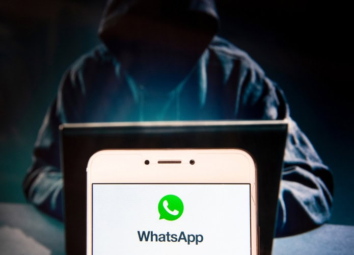 Pessoa com capuz à frente de um notebook; em primeiro plano, logo do WhatsApp em um smartphone