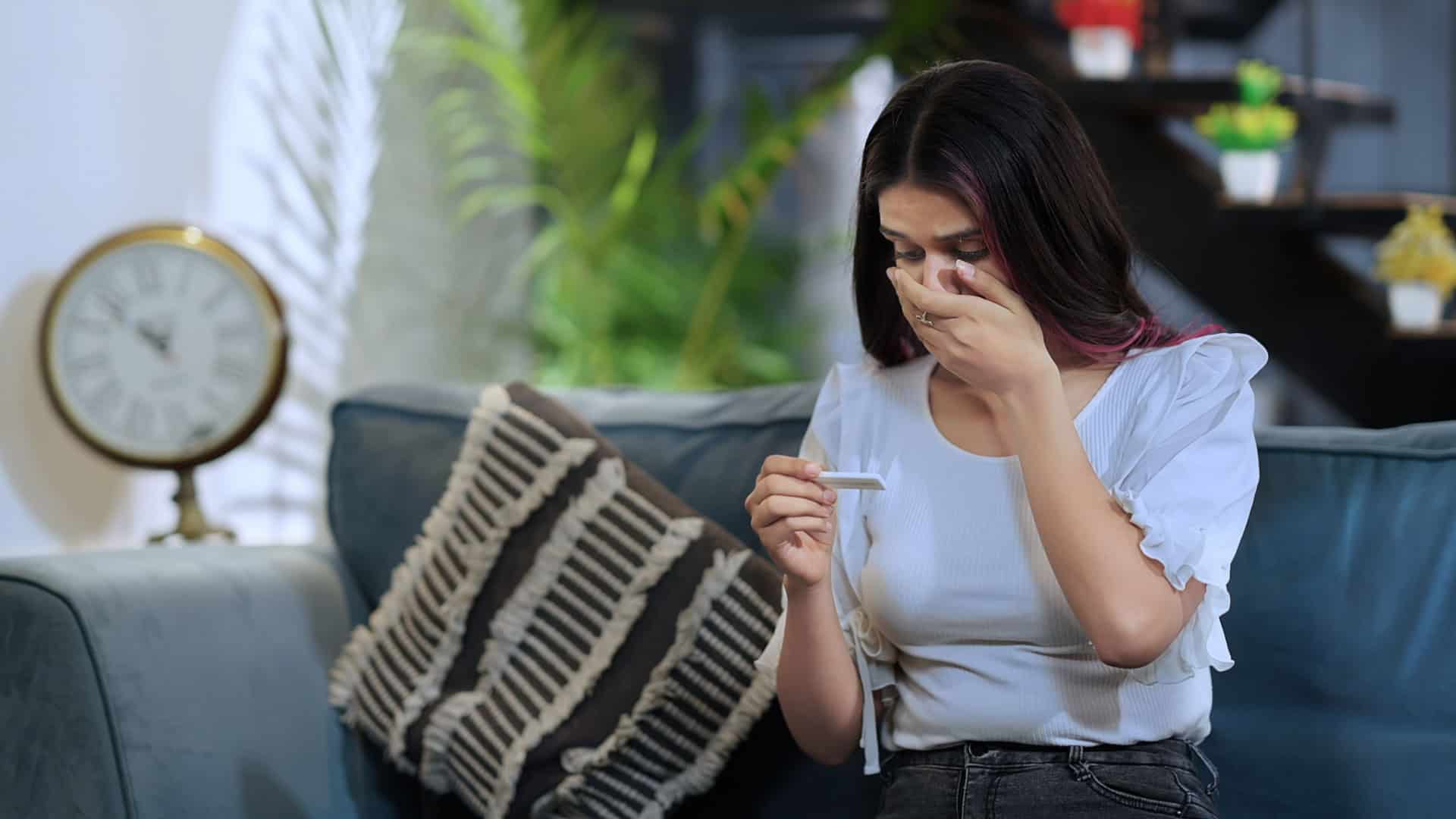 Uma pessoa sentada em um sofá segura um pequeno objeto retangular, que parece ser um teste de gravidez. Ela veste uma blusa branca com mangas franzidas e calça escura. Ao lado, há uma almofada decorativa com padrão listrado. No fundo, um grande relógio de números romanos está na parede, junto a plantas e prateleiras com vasos pequenos. A cena transmite um momento de expectativa e reflexão sobre uma possível gravidez.
