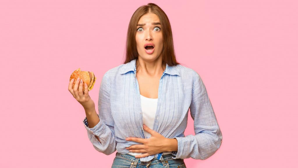 Dor de estômago. Garota segurando hambúrguer e tocando o estômago se sentindo mal depois de comer junk food em um fundo de estúdio amarelo