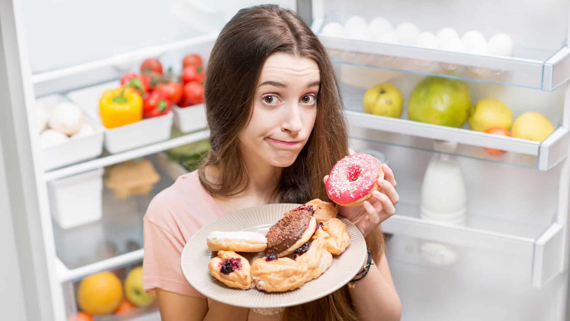 Retrato de uma jovem triste em traje de dormir com donuts doces perto da geladeira aberta