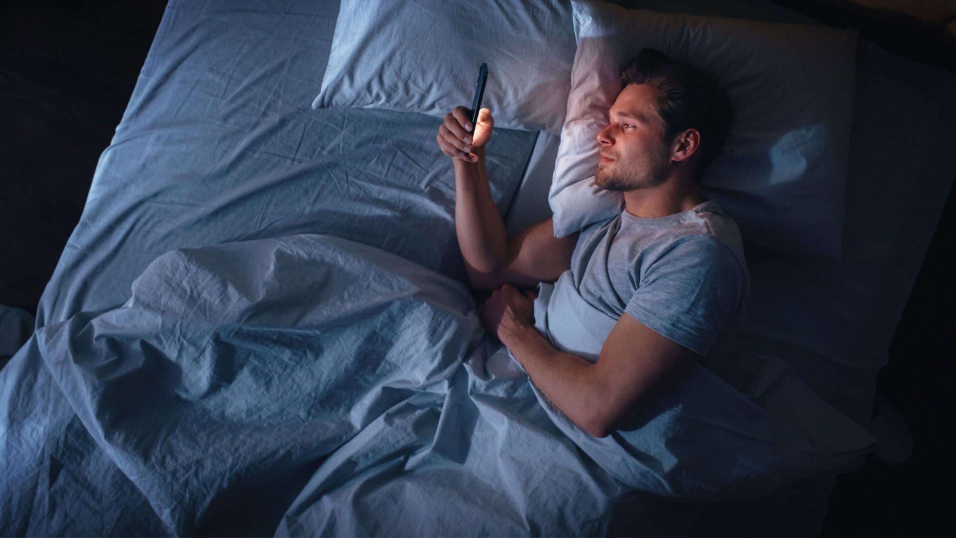 Apartamento com vista superior do quarto: jovem bonito usando smartphone na cama à noite. Cara sorridente navegando nas redes sociais, aplicativos de namoro, software de trabalho remoto, fazendo compras online na Internet no celular.