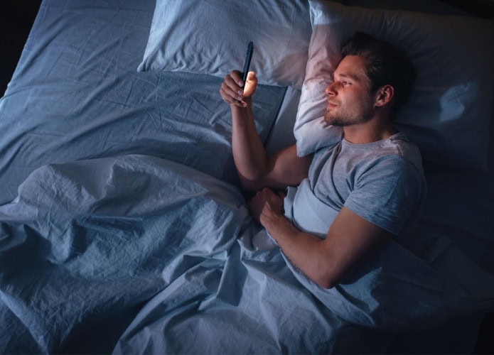 Apartamento com vista superior do quarto: jovem bonito usando smartphone na cama à noite. Cara sorridente navegando nas redes sociais, aplicativos de namoro, software de trabalho remoto, fazendo compras online na Internet no celular.