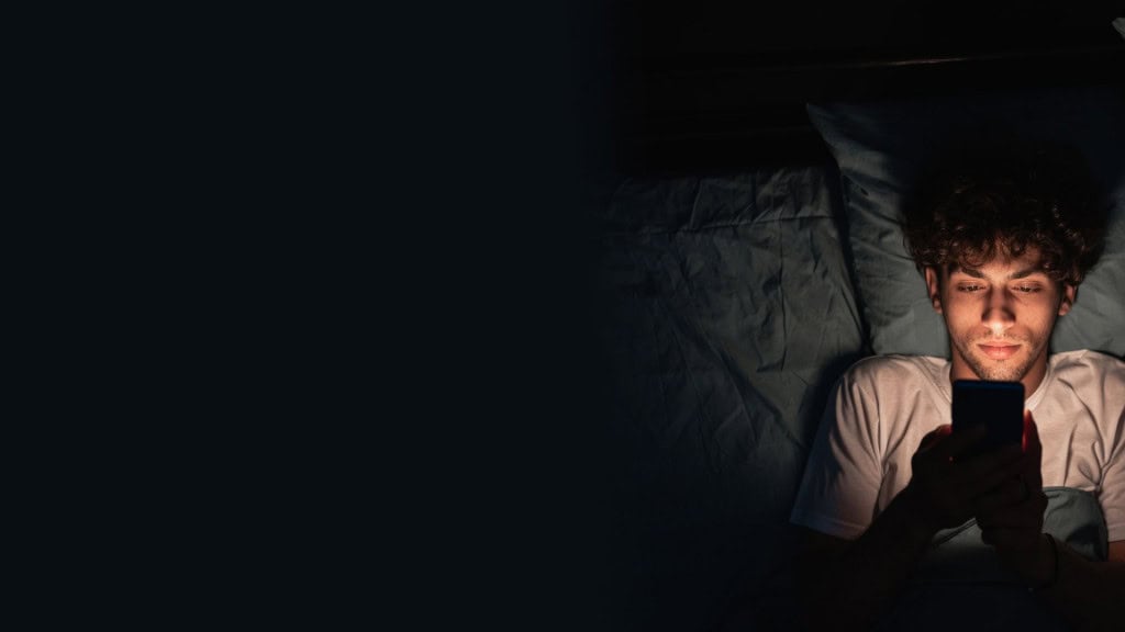 Homem jovem, sonolento e exausto deitado na cama usando smartphone tarde da noite. Insônia, conceito de distúrbio do sono. Vício em celular. 