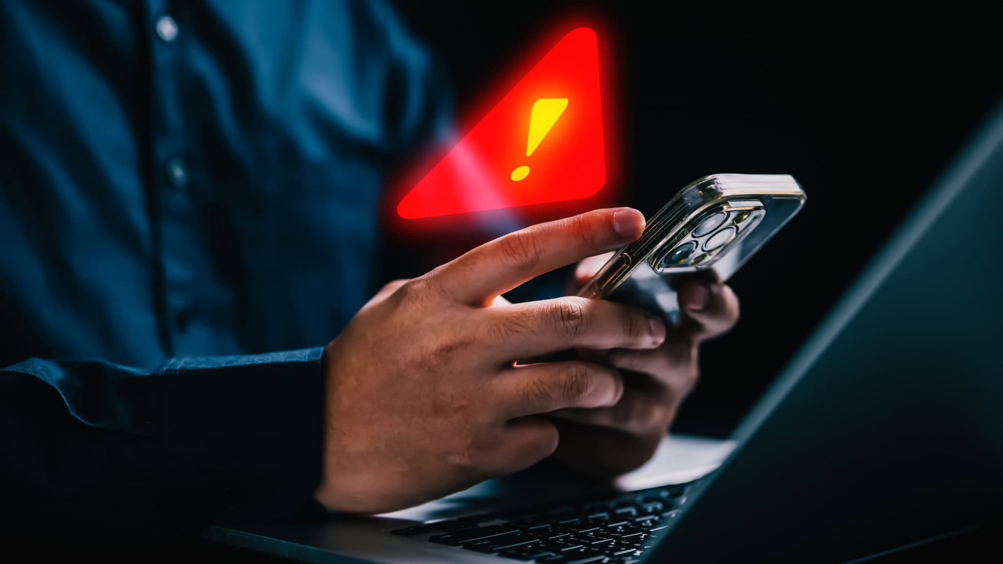 Pessoa usando iPhone em frente notebook com grande sinal de alerta holográfico em cima do celular