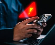 Pessoa usando iPhone em frente notebook com grande sinal de alerta holográfico em cima do celular