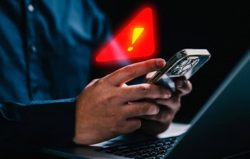 Pessoa usando iPhone em frente notebook com grande sinal de alerta holográfico em cima do celular