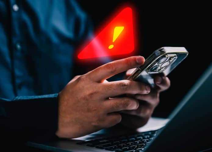 Pessoa usando iPhone em frente notebook com grande sinal de alerta holográfico em cima do celular