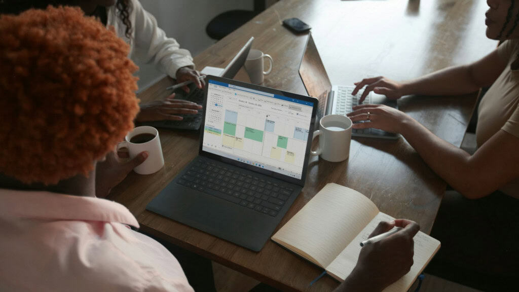 três mulheres sentadas em uma mesa, uma delas com um computador aberto usando o excel