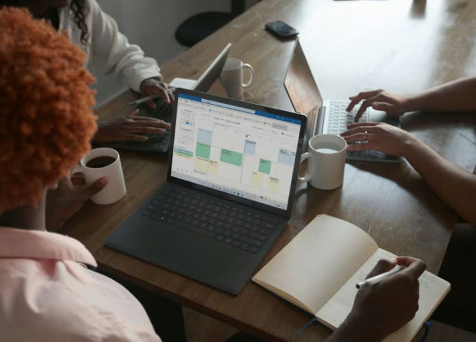 três mulheres sentadas em uma mesa, uma delas com um computador aberto usando o excel