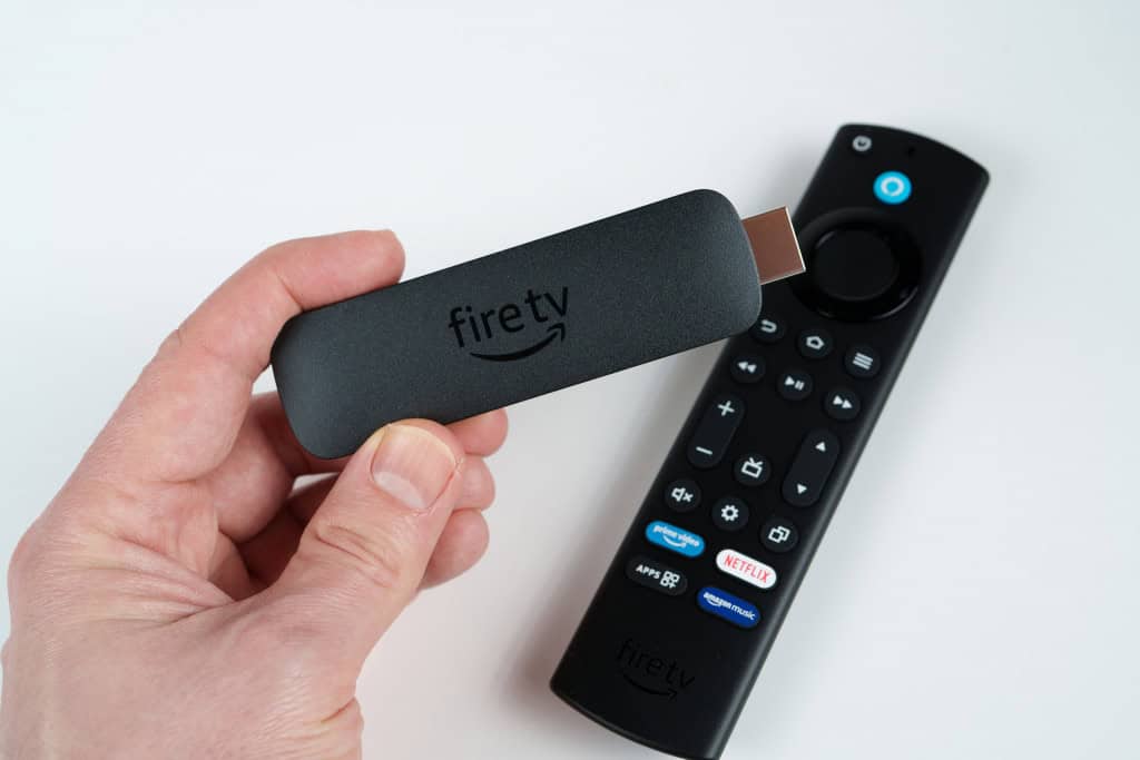 Amazon Fire TV Stick na mão de uma pessoa; mais abaixo, em uma mesa branca, o controle do aparelho