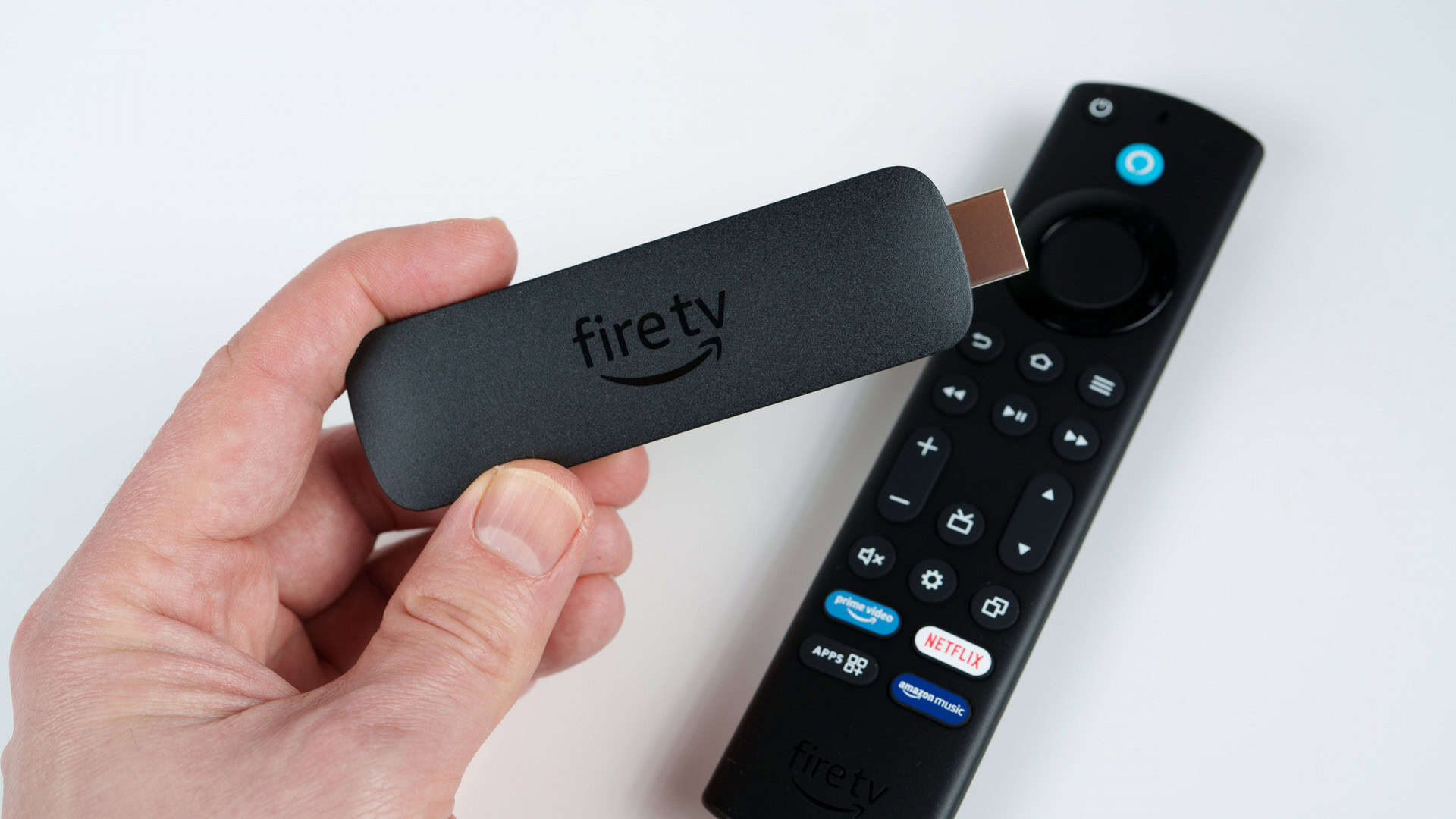 Amazon Fire TV Stick na mão de uma pessoa; mais abaixo, em uma mesa branca, o controle do aparelho