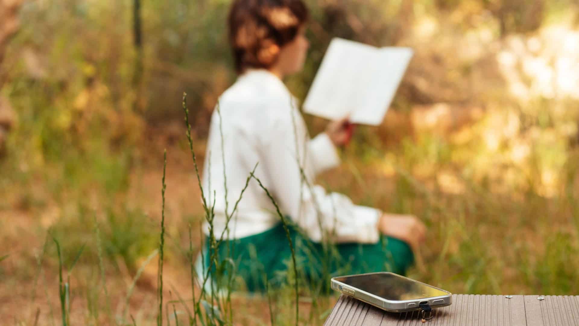 Garota lê um livro ao fundo enquanto o celular está em destaque. Representação de um “dia de silêncio” e detox de dopamina.