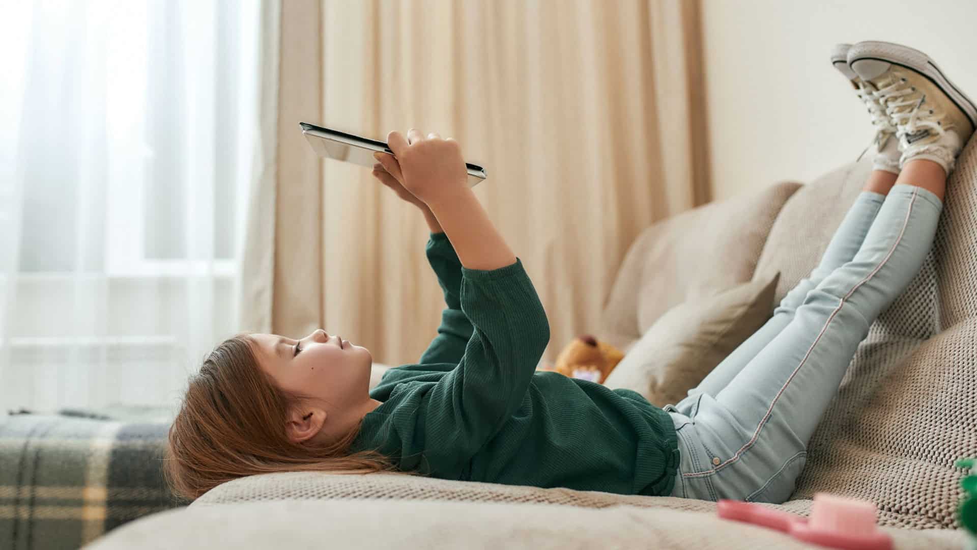 Menina pequena e fofa jogando em um tablet deitada no sofá com os pés para o alto e tênis calçados, sozinha em casa em uma sala grande. Crianças e tecnologias modernas