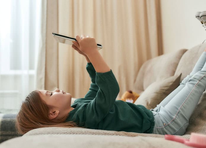 Menina pequena e fofa jogando em um tablet deitada no sofá com os pés para o alto e tênis calçados, sozinha em casa em uma sala grande. Crianças e tecnologias modernas