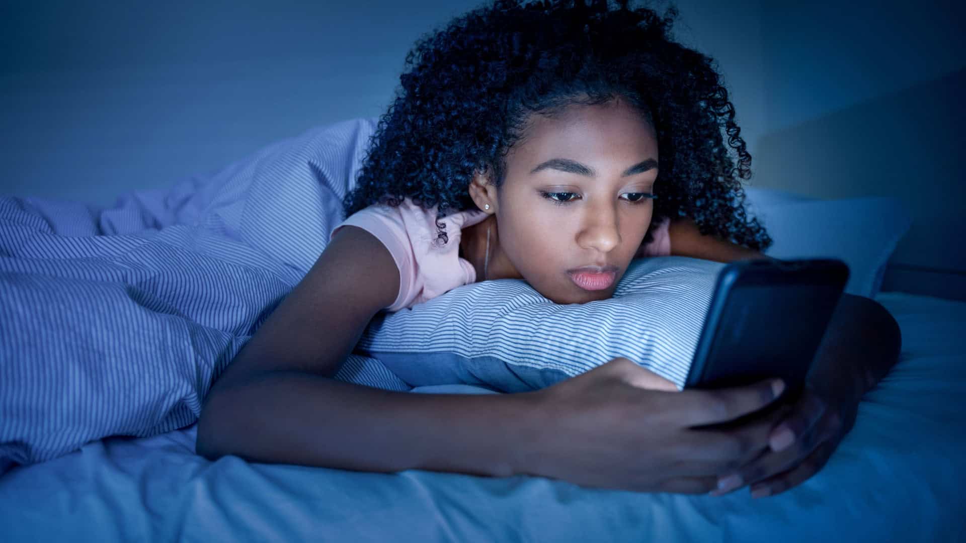 Garota negra usando o smartphone na cama antes de dormir. / Crédito: F01 PHOTO (Shutterstock/reprodução)