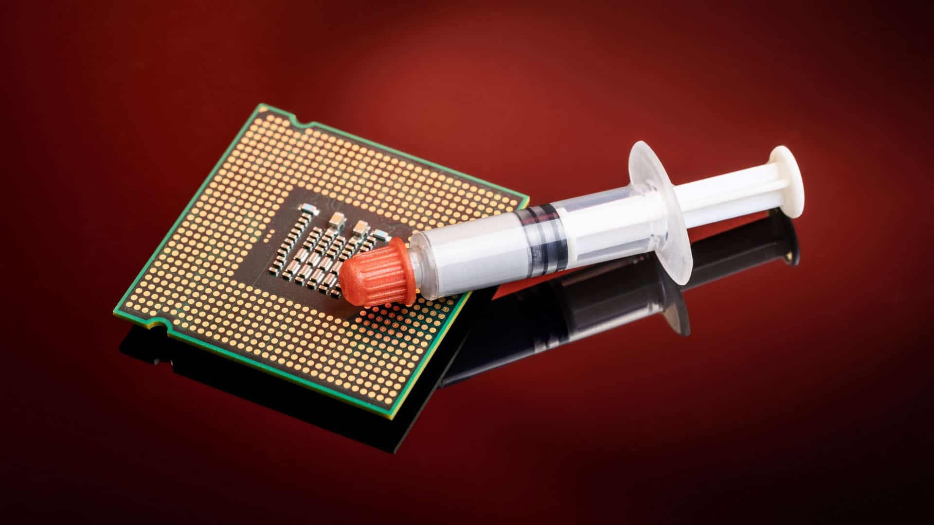 imagem mostra uma mesa de metal vermelha. em cima dela há um grande chip e em cima deste há uma seringa lacrada com um conteúdo branco dentro (pasta térmica)