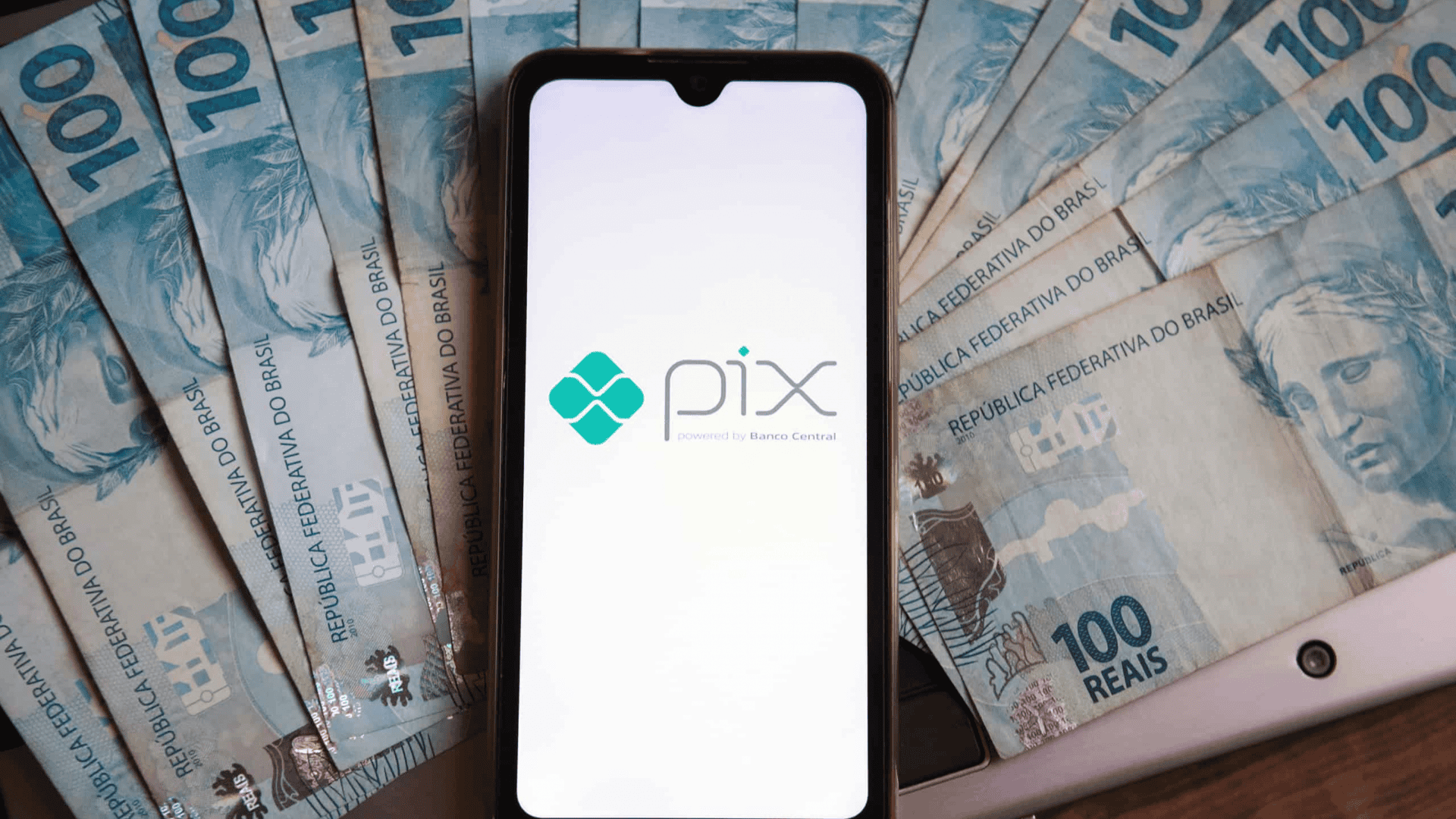 Pessoa com a função do PIX sendo iniciada no celular e várias notas de R$ 100 ao redor do aparelho