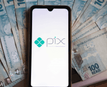 Pessoa com a função do PIX sendo iniciada no celular e várias notas de R$ 100 ao redor do aparelho