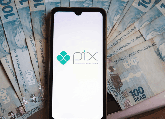 Pessoa com a função do PIX sendo iniciada no celular e várias notas de R$ 100 ao redor do aparelho