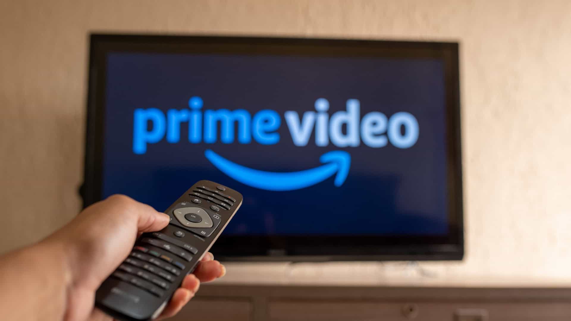 Logo do Prime Video em uma TV ao fundo, desfocada; à frente, uma pessoa com um controle remoto de TV na mão