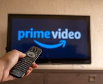 Logo do Prime Video em uma TV ao fundo, desfocada; à frente, uma pessoa com um controle remoto de TV na mão