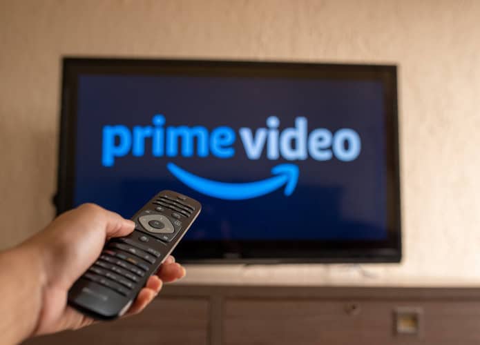 Logo do Prime Video em uma TV ao fundo, desfocada; à frente, uma pessoa com um controle remoto de TV na mão