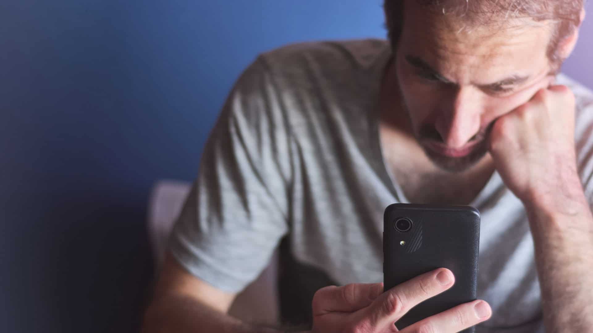 Homem caucasiano de 30 anos sentado em casa olhando para a tela do celular com expressão entediada e desinteressada, em frente a uma parede azul. Tédio, vício em tecnologia, isolamento e depressão. / Crédito: Edaccor (Shutterstock/reprodução)