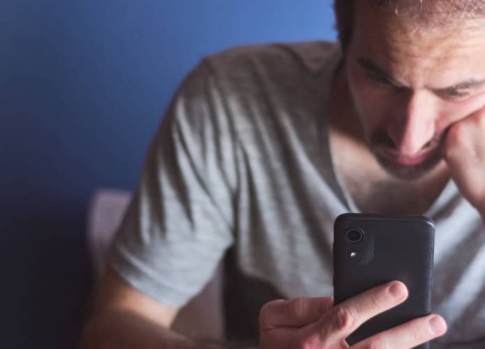Homem caucasiano de 30 anos sentado em casa olhando para a tela do celular com expressão entediada e desinteressada, em frente a uma parede azul. Tédio, vício em tecnologia, isolamento e depressão. / Crédito: Edaccor (Shutterstock/reprodução)