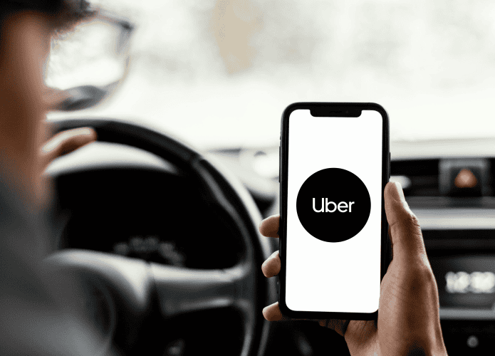Motorista segura celular com o app da Uber aberto enquanto está ao volante, com interior do carro desfocado ao fundo