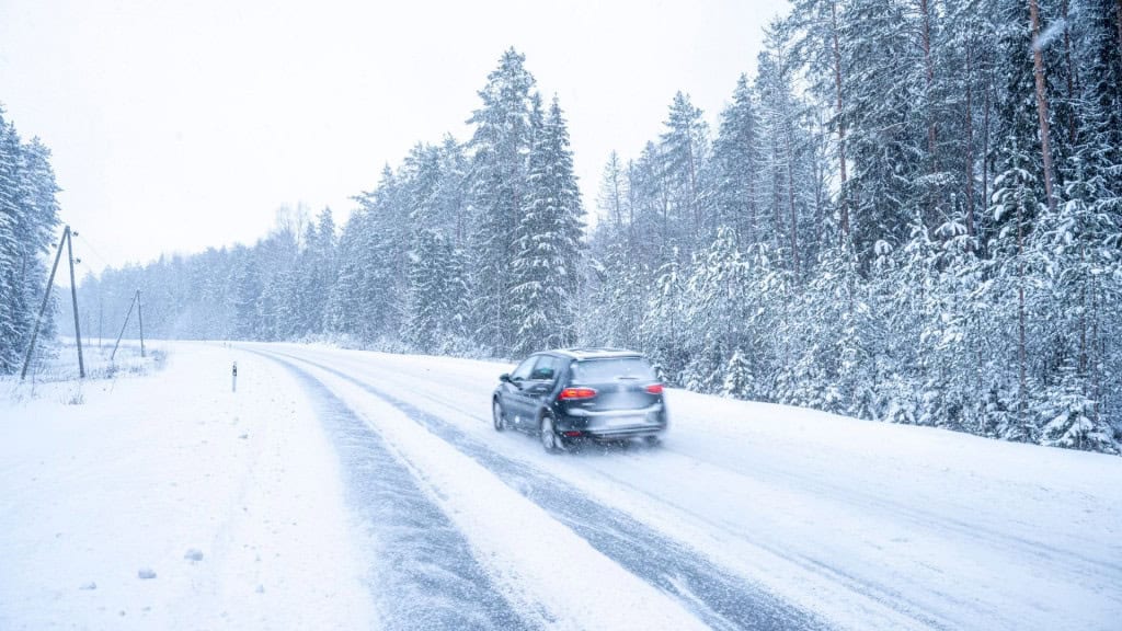 Um carro trafega por uma estrada coberta de neve, ladeada por árvores altas igualmente nevadas. A paisagem transmite isolamento e atenção, típica de viagens em regiões frias e desafiadoras.