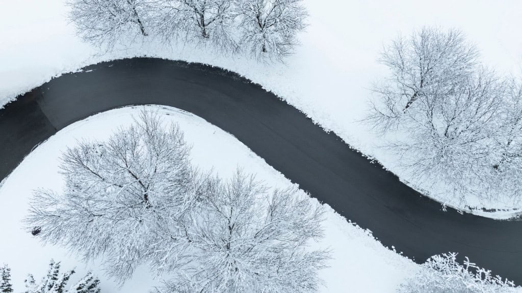 Uma estrada sinuosa em formato de “S” atravessa uma paisagem inteiramente coberta de neve, vista de cima. As árvores ao redor estão carregadas de branco, criando um contraste marcante com o asfalto escuro e a curva elegante da via.