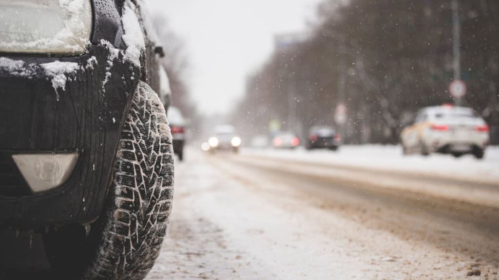 Um carro preto está parado com neve acumulada sobre o capô e os pneus parcialmente cobertos de gelo. Ao fundo, vê-se uma estrada também tomada pela neve, com outros veículos distantes sob o céu nublado.
