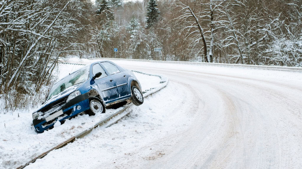 Um carro azul está fora da estrada em uma curva coberta de neve, com a frente danificada. A cena sugere um acidente causado por pista escorregadia em condições de inverno rigoroso.