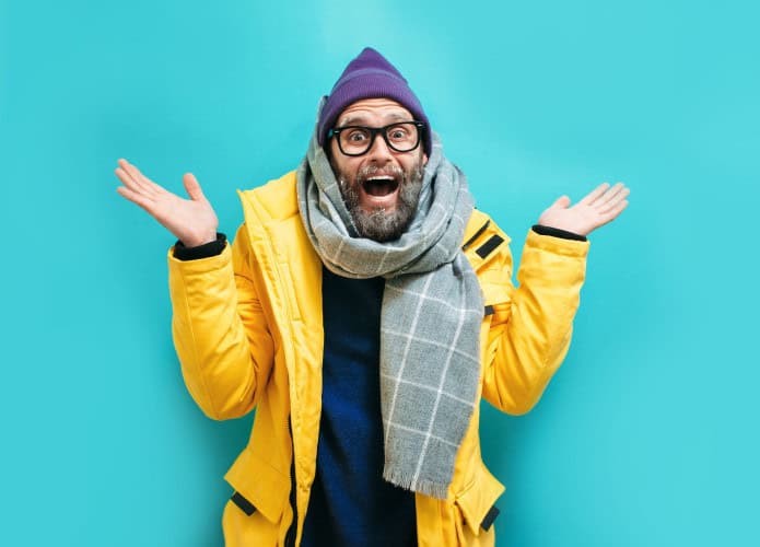 🧥 A imagem mostra uma pessoa em pé diante de um fundo azul vibrante. Ela usa jaqueta amarela, suéter azul escuro, cachecol xadrez cinza e gorro roxo. Com os braços levantados e as palmas voltadas para cima, seu gesto parece expressar dúvida ou surpresa.