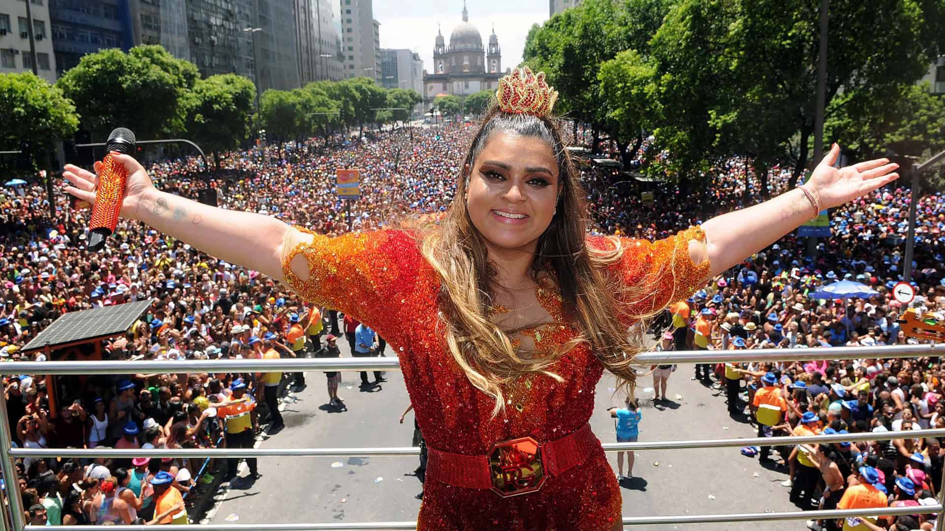Uma multidão animada ocupa uma avenida larga no centro do Rio de Janeiro durante o desfile de um bloco de carnaval. No centro da imagem, a cantora Preta Gil aparece sorridente sobre um trio elétrico, vestida com fantasia brilhante, segurando um microfone. Ela interage com o público, que está vestido com roupas coloridas e adereços carnavalescos. Ao fundo, prédios altos em estilo antigo contrastam com o céu azul. A atmosfera é festiva e vibrante, com pessoas dançando, cantando e celebrando juntas.