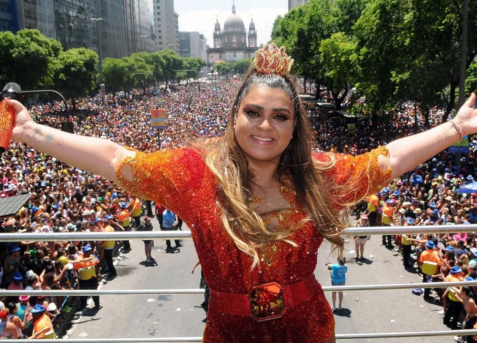 Uma multidão animada ocupa uma avenida larga no centro do Rio de Janeiro durante o desfile de um bloco de carnaval. No centro da imagem, a cantora Preta Gil aparece sorridente sobre um trio elétrico, vestida com fantasia brilhante, segurando um microfone. Ela interage com o público, que está vestido com roupas coloridas e adereços carnavalescos. Ao fundo, prédios altos em estilo antigo contrastam com o céu azul. A atmosfera é festiva e vibrante, com pessoas dançando, cantando e celebrando juntas.