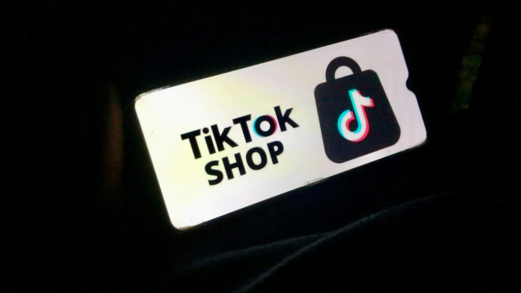 A imagem mostra um celular exibindo o logo e o texto "TikTok Shop" em uma tela branca, destacando-se de um fundo escuro. Basicamente, é uma representação visual do TikTok Shop, a funcionalidade de comércio eletrônico do TikTok. O TikTok Shop foi lançado no Brasil em maio de 2025 e permite que os usuários comprem produtos diretamente no aplicativo, através de vídeos, lives e perfis de lojas, com influenciadores sendo comissionados pelas vendas.