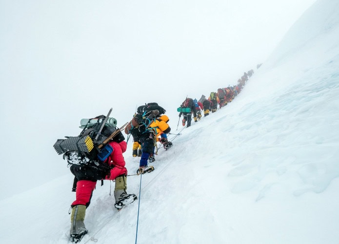 Uma longa fila de alpinistas sobe uma encosta íngreme e coberta de neve, conectados por cordas para segurança. Cada pessoa carrega mochilas pesadas e equipamentos de escalada, avançando com esforço em meio ao gelo e altitude extrema. A imagem transmite trabalho em equipe, superação e os desafios intensos do montanhismo em grandes altitudes.
