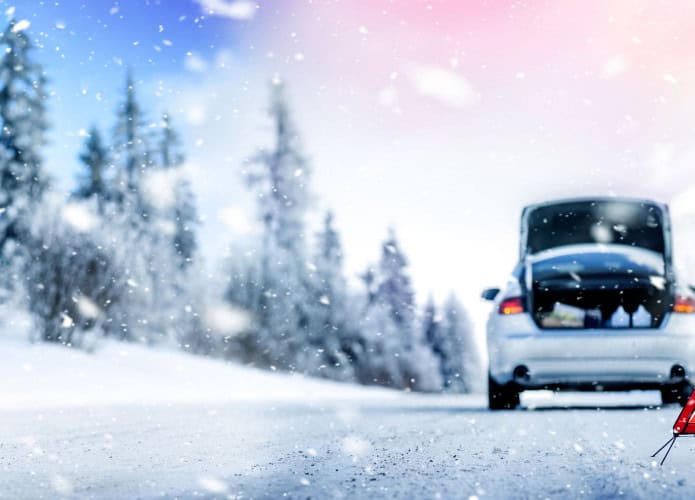 Um carro está parado em uma estrada nevada com o porta-malas aberto, sugerindo uma situação de emergência. Um triângulo de sinalização está posicionado atrás do veículo, alertando outros motoristas sobre o ocorrido.