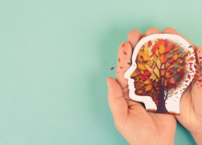 Duas mãos seguram um recorte em forma de cabeça humana, com uma árvore colorida dentro, cujas folhas parecem se desprender. O fundo verde claro destaca a ideia de memória se esvaindo, simbolizando o impacto do Alzheimer.