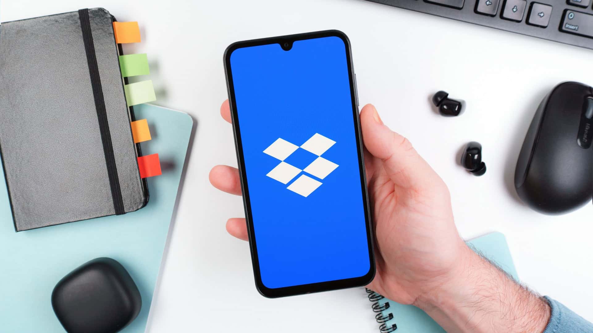Pessoa segurando celular exibindo logotipo do Dropbox; pessoa segura celular em cima de mesa de escritório com caderno, fones de ouvido, mouse e teclado ao fundo
