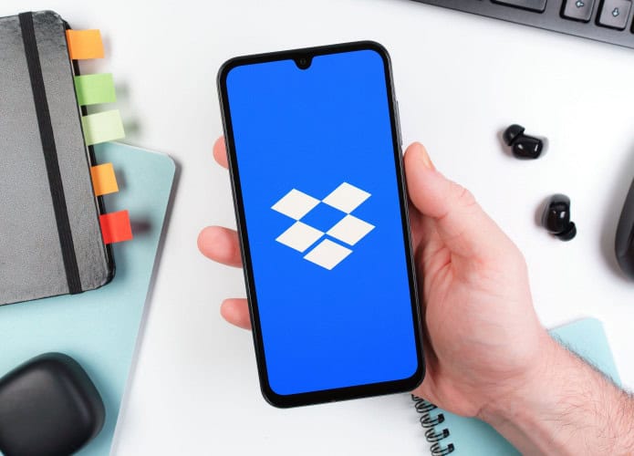 Pessoa segurando celular exibindo logotipo do Dropbox; pessoa segura celular em cima de mesa de escritório com caderno, fones de ouvido, mouse e teclado ao fundo