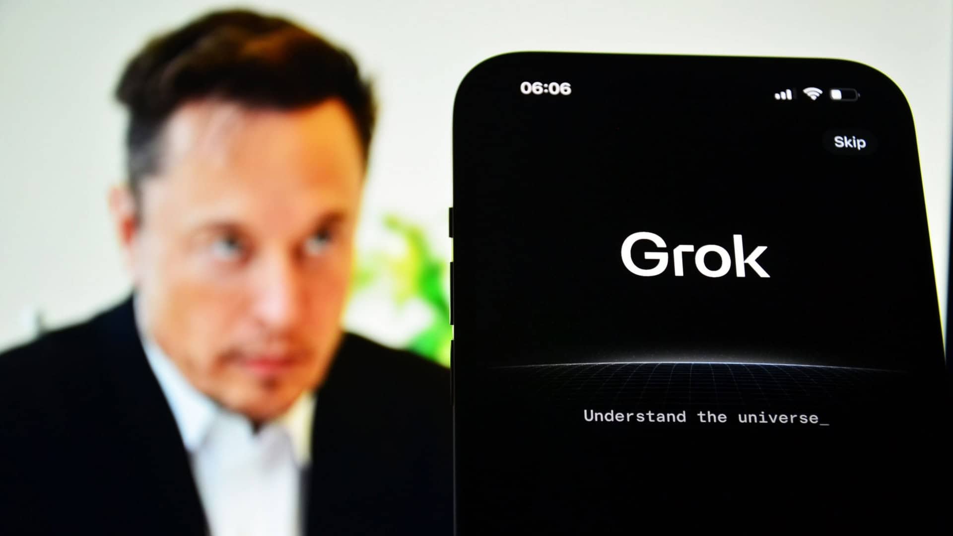 Celular exibindo página sobre Grok colocado na frente de tela exibindo foto de Elon Musk