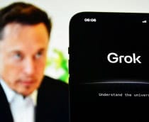 Celular exibindo página sobre Grok colocado na frente de tela exibindo foto de Elon Musk