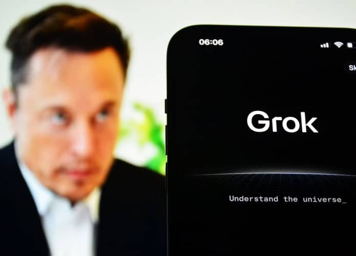 Celular exibindo página sobre Grok colocado na frente de tela exibindo foto de Elon Musk