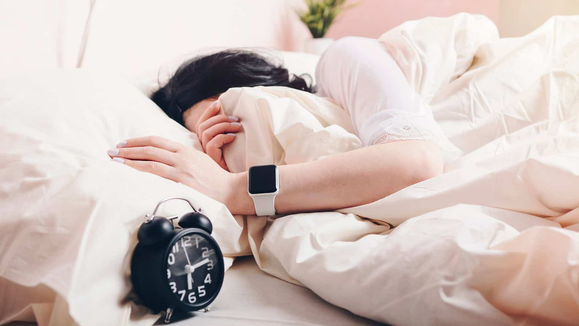 Mulher usando Apple Watch enquanto dorme enterrada nas cobertas ao lado de um relógio despertador