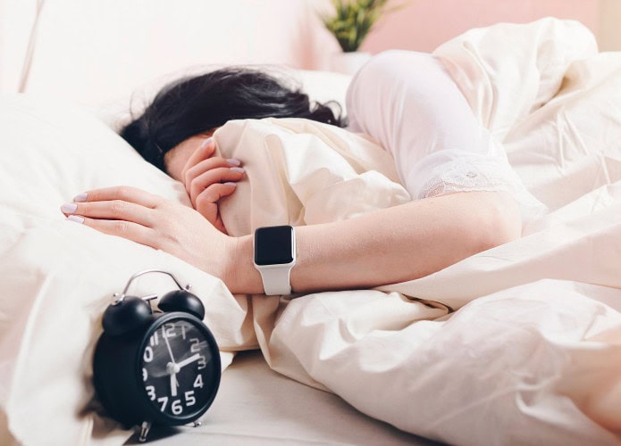 Mulher usando Apple Watch enquanto dorme enterrada nas cobertas ao lado de um relógio despertador