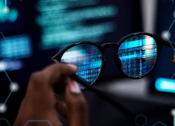 Montagem com pessoa segurando óculos de grau na frente de computador; nas lentes, aparecem códigos binários