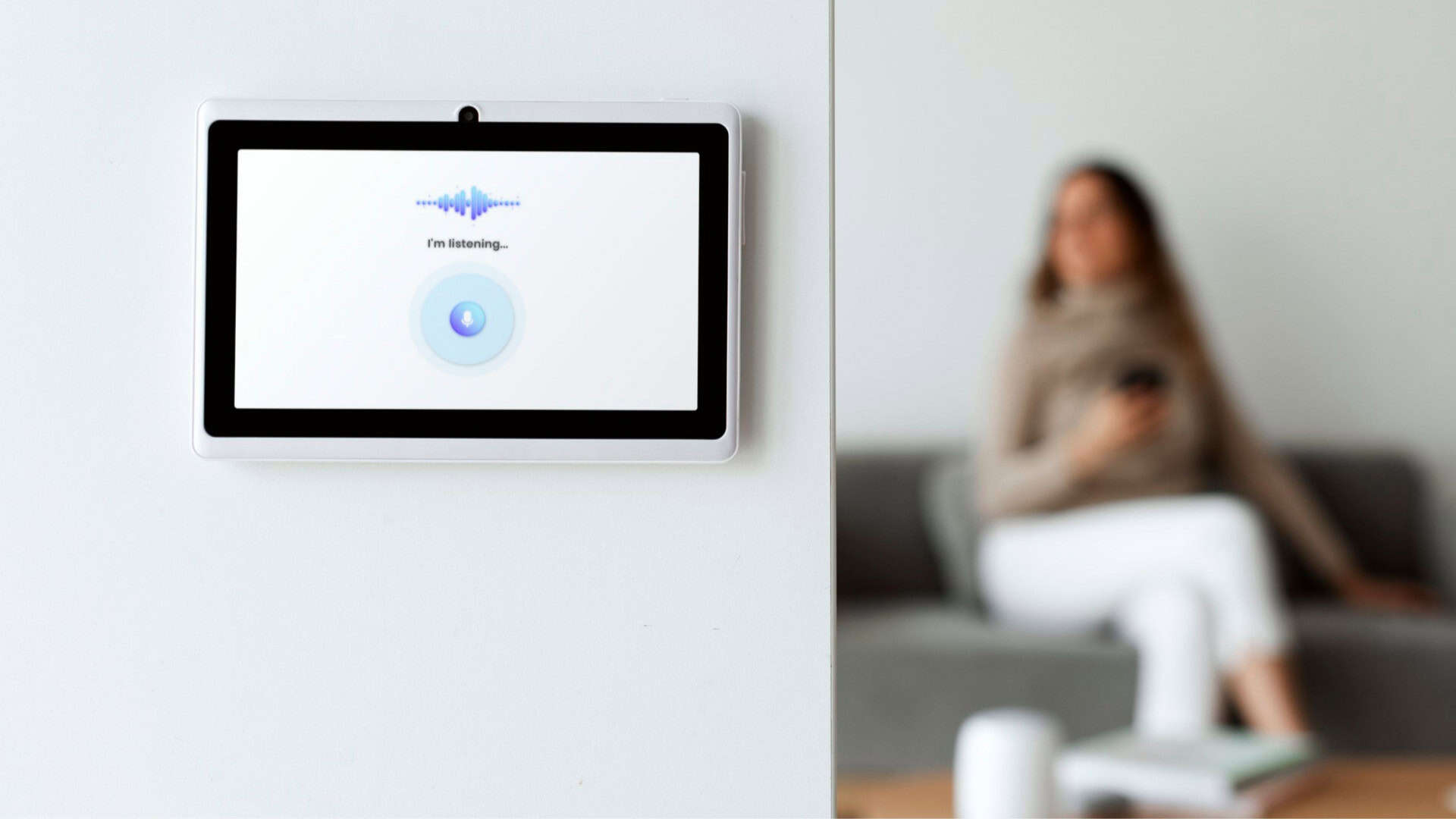 visão em primeiro plano de um monitor de casa inteligente, com mulher sentada em sofá em segundo plano, desfocada