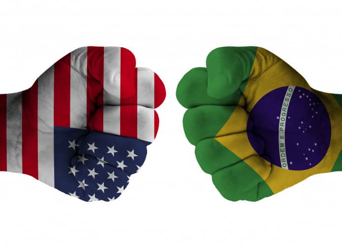 Duas mãos com punhos cerrados e uma apontando para a outra; a da esquerda está com a bandeira dos EUA desenhada; a da direita tem o desenho da bandeira do Brasil